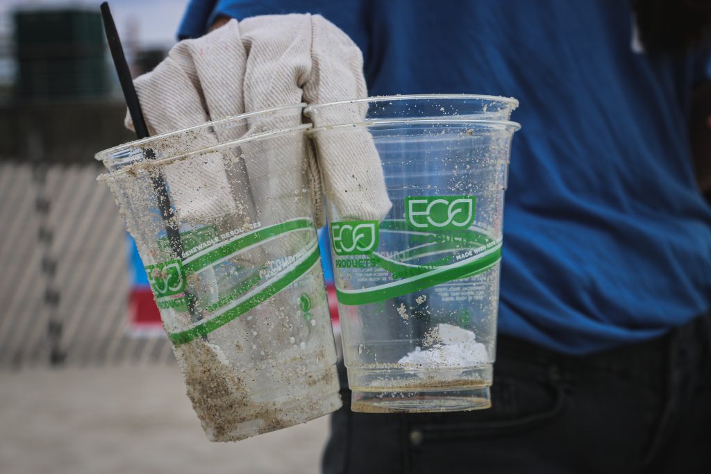 Fotografia de uma mão, com luvas de coleta de lixo, segurando dois copos plásticos cobertos de areia da praia. Nos copos, lê-se "eco" e há grafismos verdes que remetem à sustentabilidade.