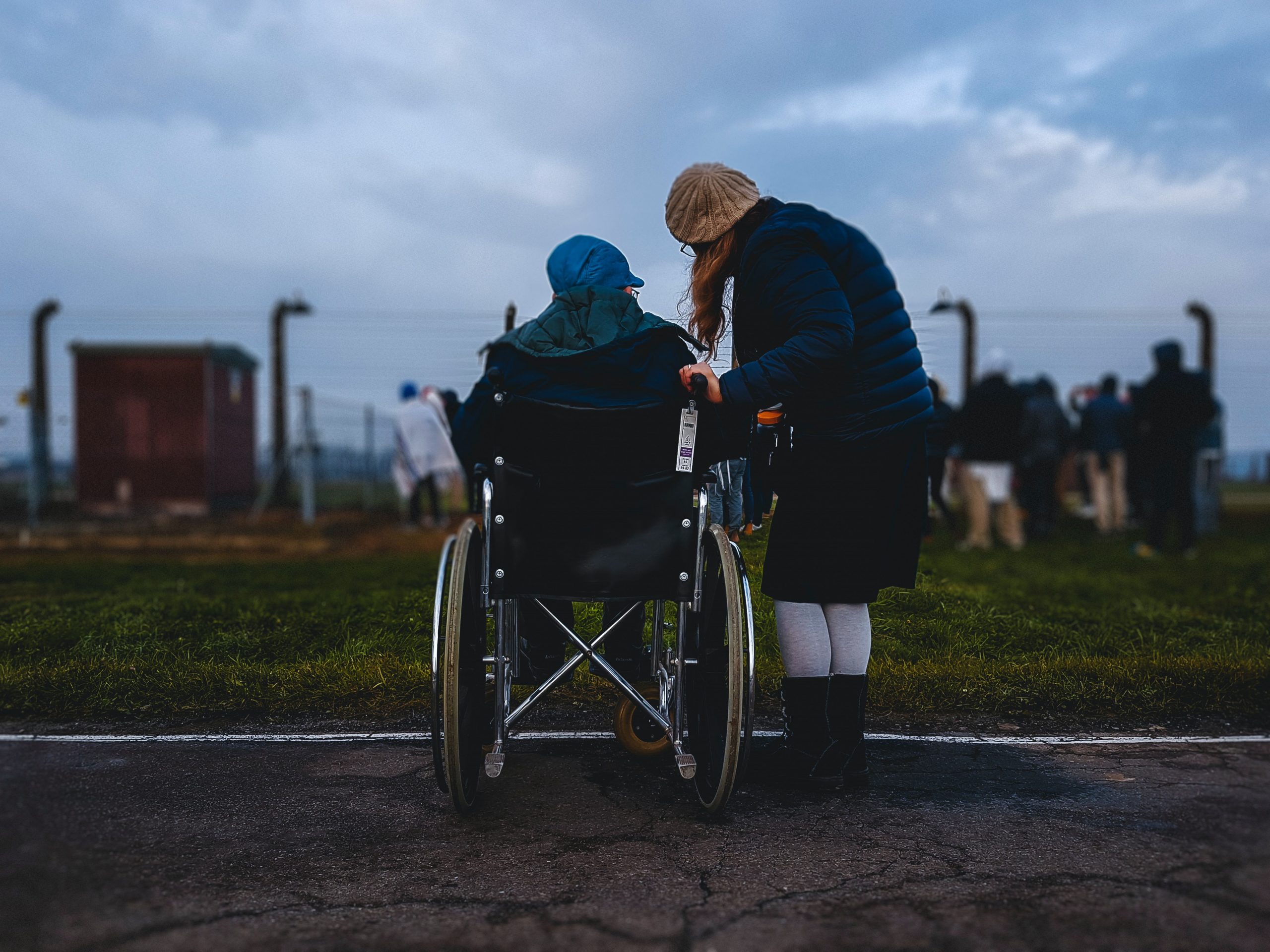 Fotografia de duas pessoas de costas. Uma é uma pessoa com deficiência, em uma cadeira de rodas, e outra é uma pessoa sem deficiência, ao lado dela. Ambas olham para o horizonte.