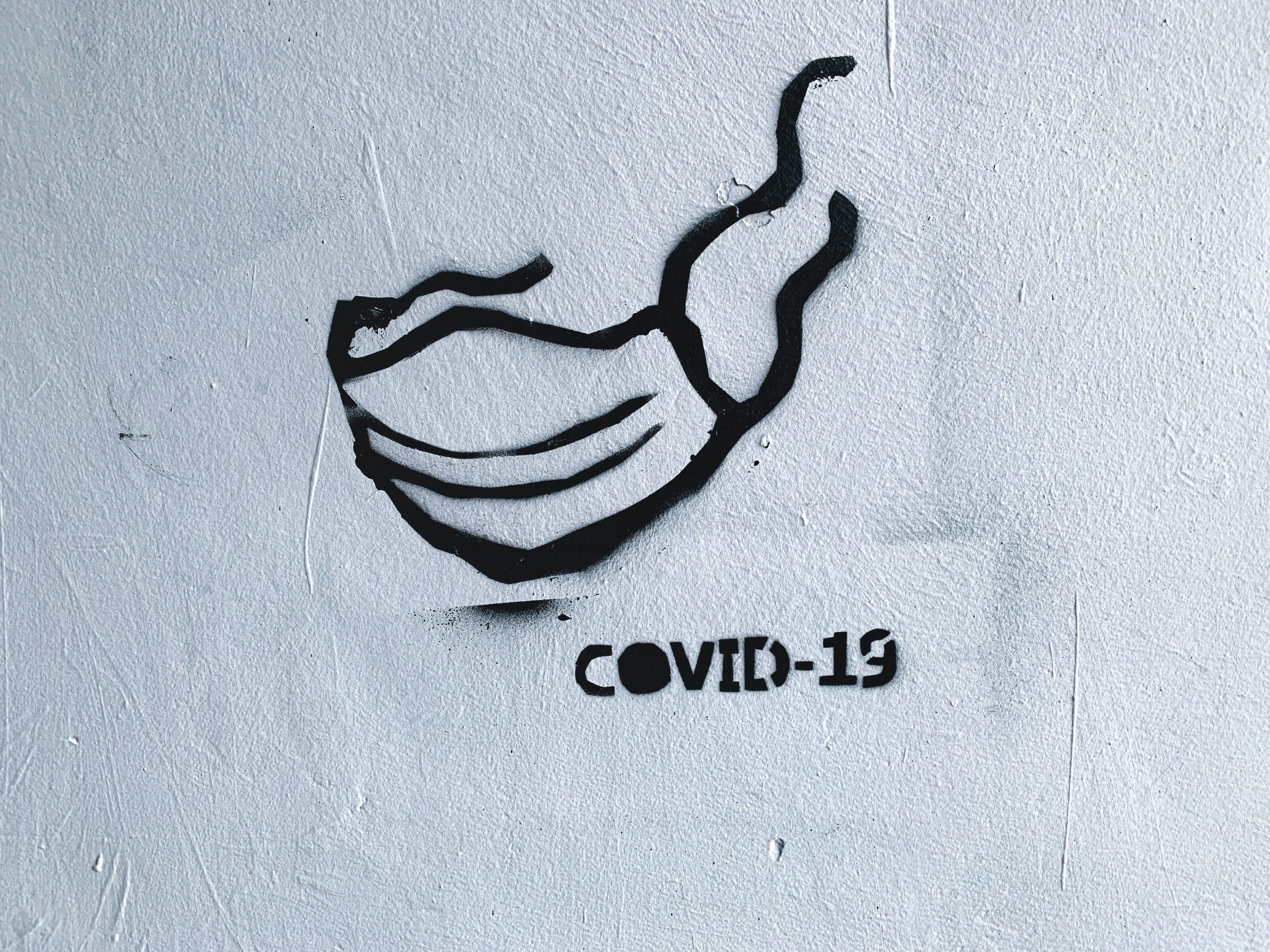 Fotografia de uma parede branca na qual se encontra uma pichação de uma máscara cirúrgica e a palavra covid-19.