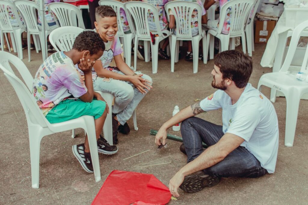 Fotografia de uma ação de voluntariado em que um homem adulto, sentado no chão, conversando com dois meninos que estão sentados em cadeiras ilustra pesquisa do Atados sobre voluntariado.