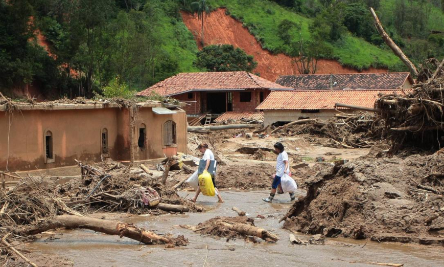 Fotografia da cidade de Petrópolis após as fortes chuvas. Algumas casas aparecem ao fundo destruídas e com muita lama, enquanto duas mulheres atravessam a lama com capas de chuva e sacolas com doações.