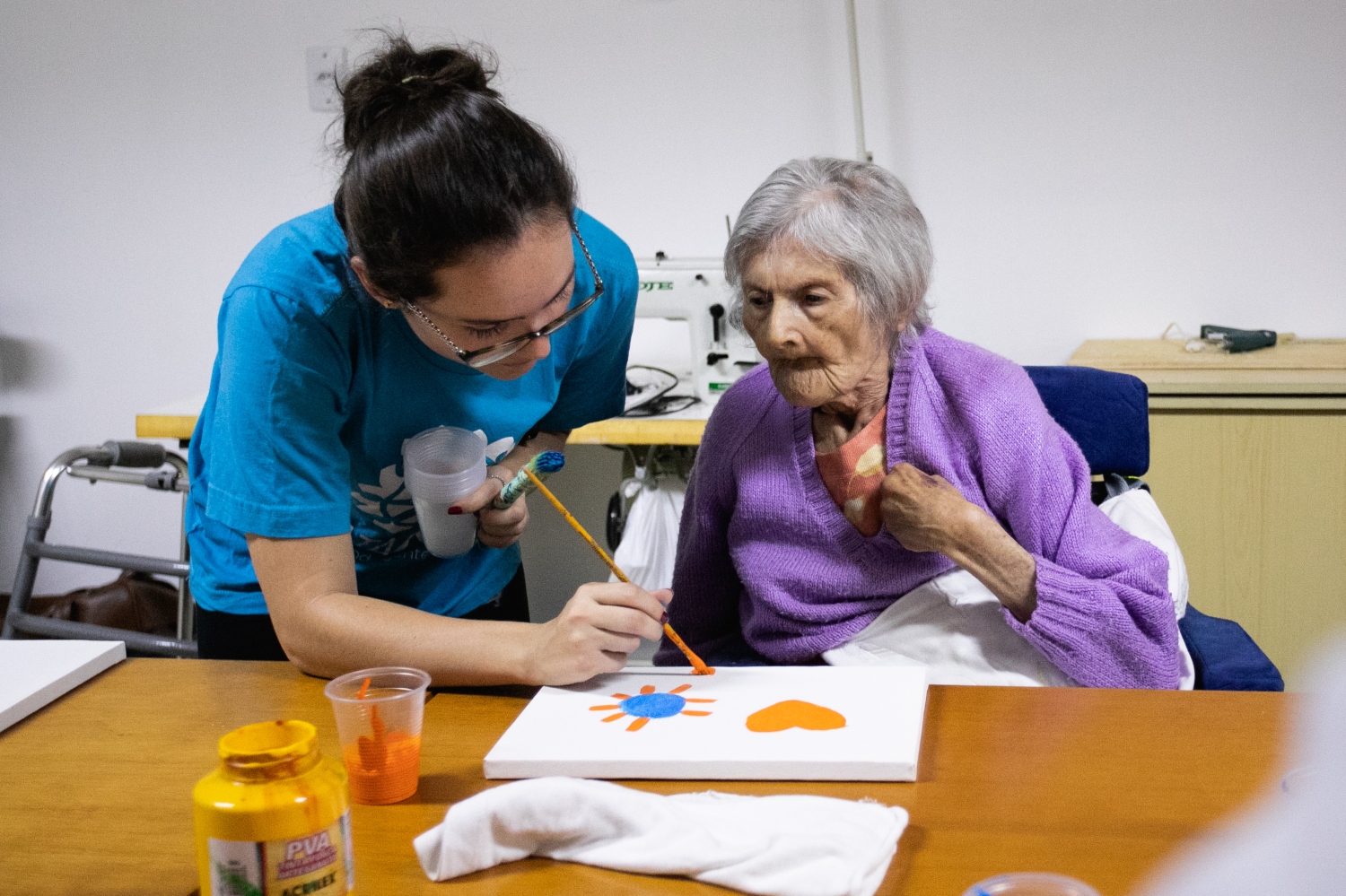 Fotografia de uma voluntária com uma senhora idosa. A voluntária ajuda a senhora a pintar um quadro colorido.